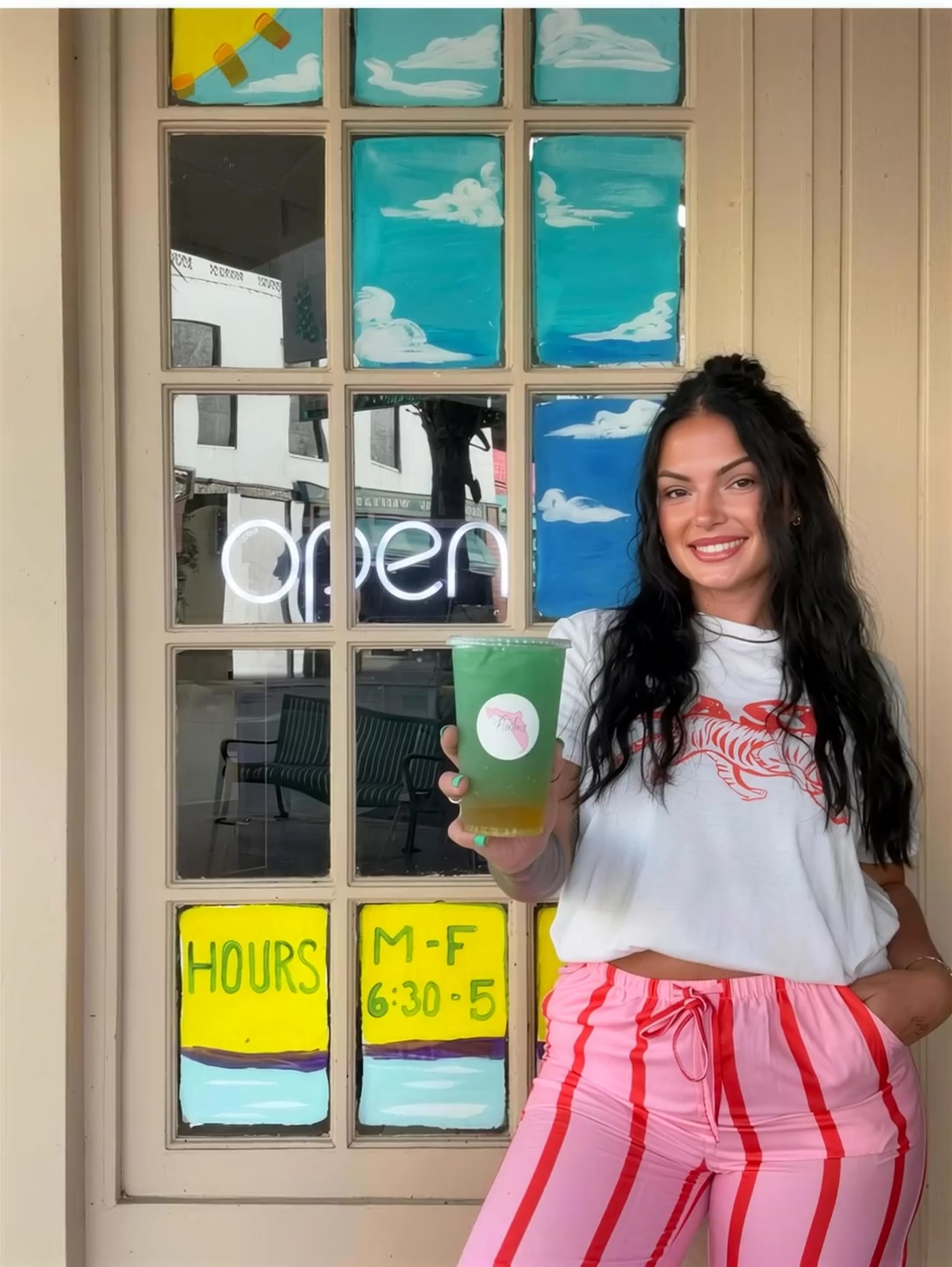 The owner of Riverfront Nutrition at the shop door with a cold green loaded tea, in front of the hand-painted windows and the neon open sign.